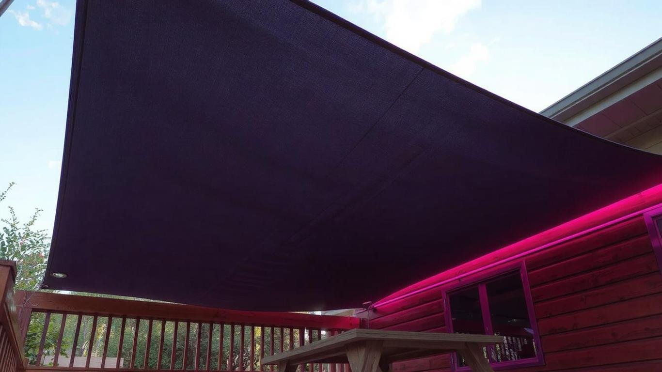 A rectangular shade sail in dark grey, hung over a wooden deck. The tarp has stainless steel D-ring grommets at the corners, secured with adjustable ropes. The scene includes a picnic table and a clear sky, highlighting the shade provided by the tarp.