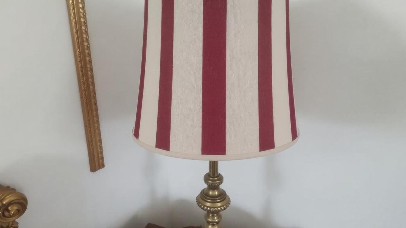 A vintage-style lamp shade with vertical stripes in cream and burgundy, paired with a brass base, placed on an ornate bedside table.