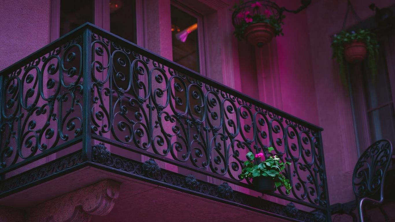 A classic wrought iron balcony railing with intricate floral and vine patterns, painted matte black. The setting is a vintage-style apartment building, complemented by hanging flower pots and antique outdoor furniture.